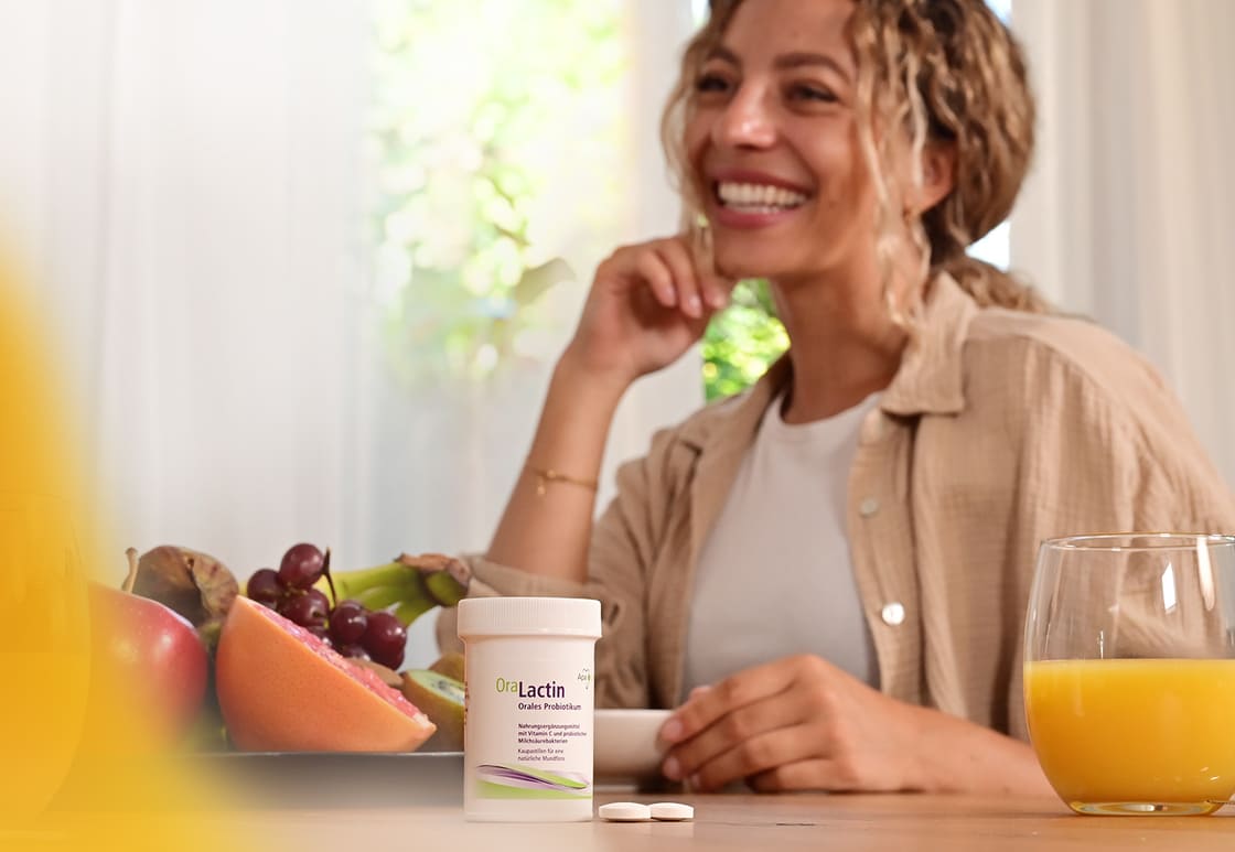 OraLactin Kaupastillen stehen auf Tisch mit Obst und Orangnsaft, im Hintergrund sitzt eine Frau